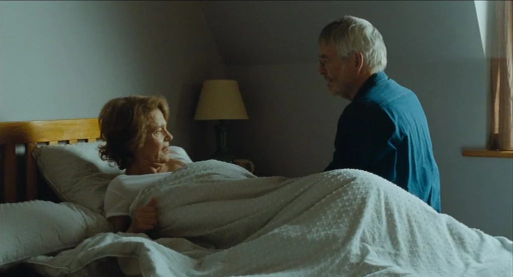 45 Years (2015) movie still: medium — A bedroom contains a wooden bed frame, a bedside lamp, and a window. A man sits beside a woman who is lying in…