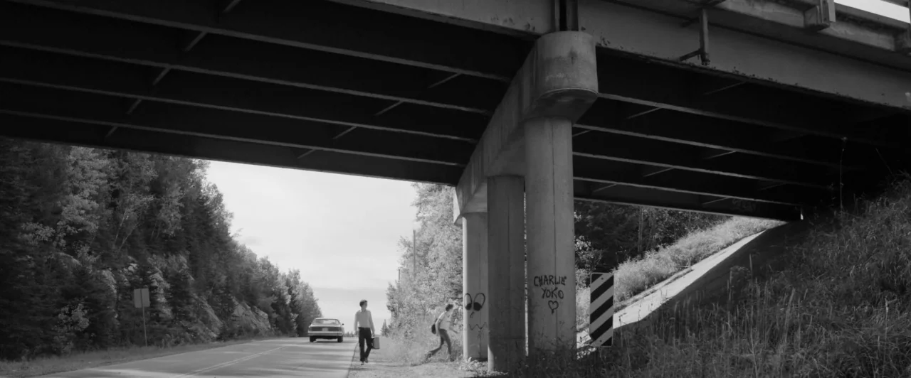 Weirdos (2016) movie still: wide — A highway passes underneath a large concrete bridge with graffiti on the support pillars. Two men walk along the…