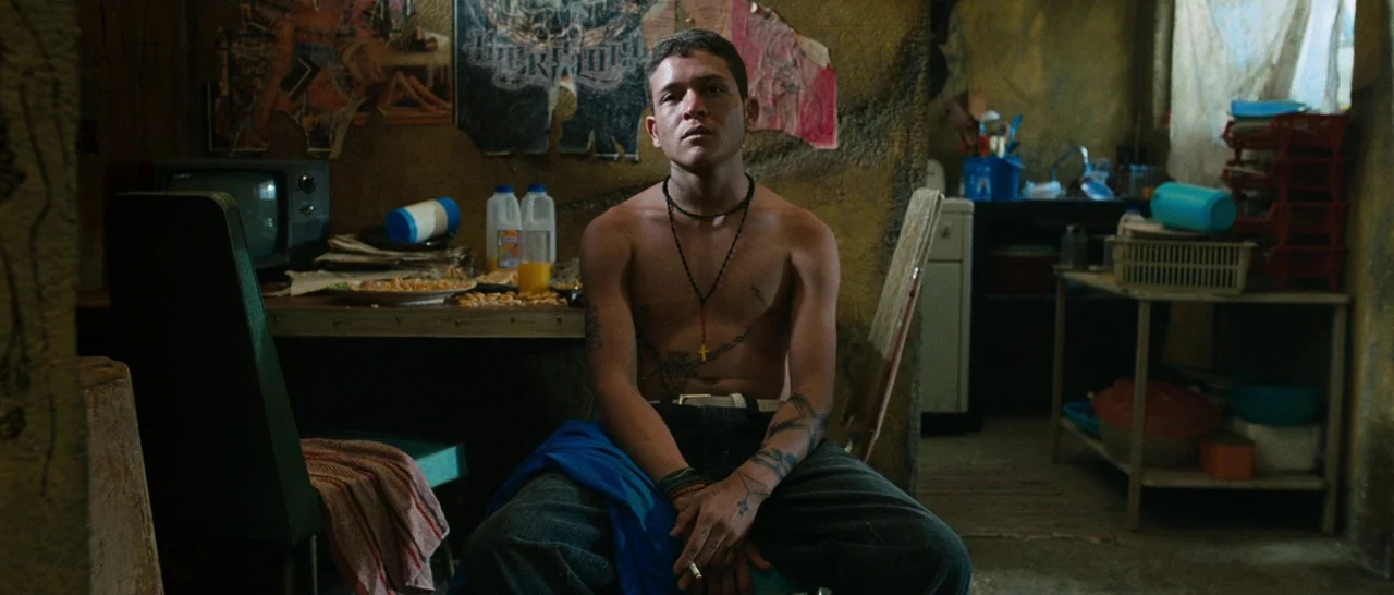 Sin Nombre (2009) movie still: medium — A cluttered, small room with textured walls decorated with posters and sparse furniture. A shirtless young…