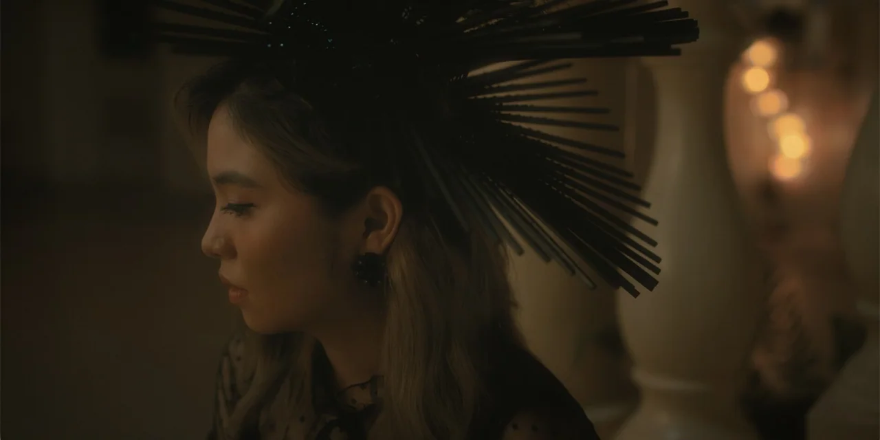 Ten Little Mistresses (2023) movie still: closeup — A dimly lit interior space features a blurred background with soft golden light points. A young…