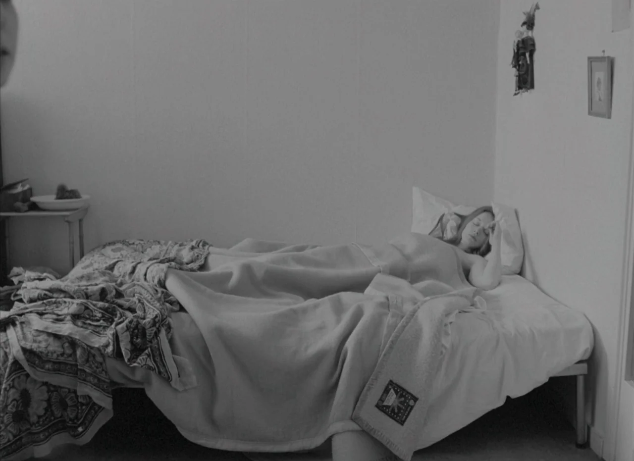 Je Tu Il Elle (1974) movie still: medium — A spartan bedroom features a single bed against a plain white wall. A young woman lies resting beneath a…