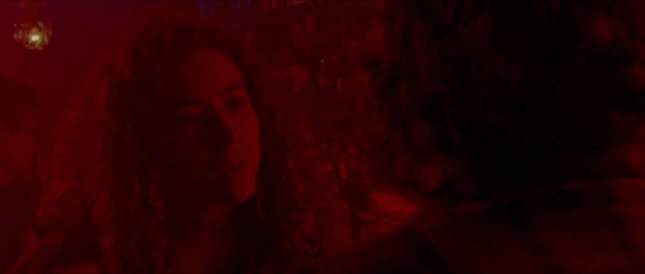 Bliss movie still: medium — A dimly lit bar is flooded with intense monochromatic red light. A young woman with curly hair looks toward someone out…