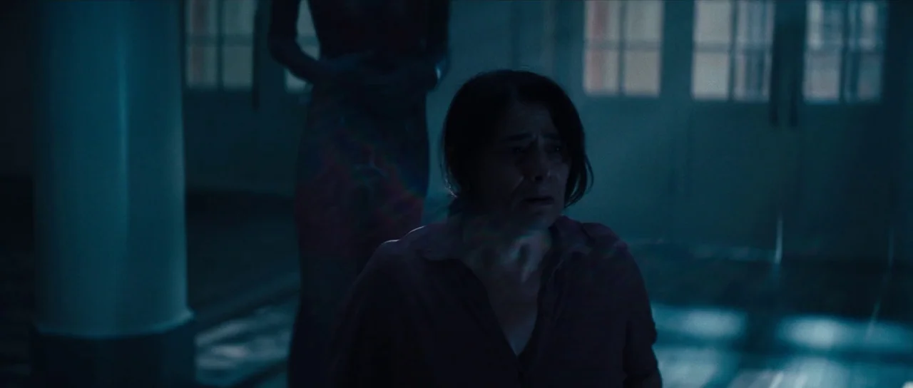 Hellraiser (2022) movie still: medium — A dimly lit indoor space featuring a hallway with white columns and distant windows. A woman in the…