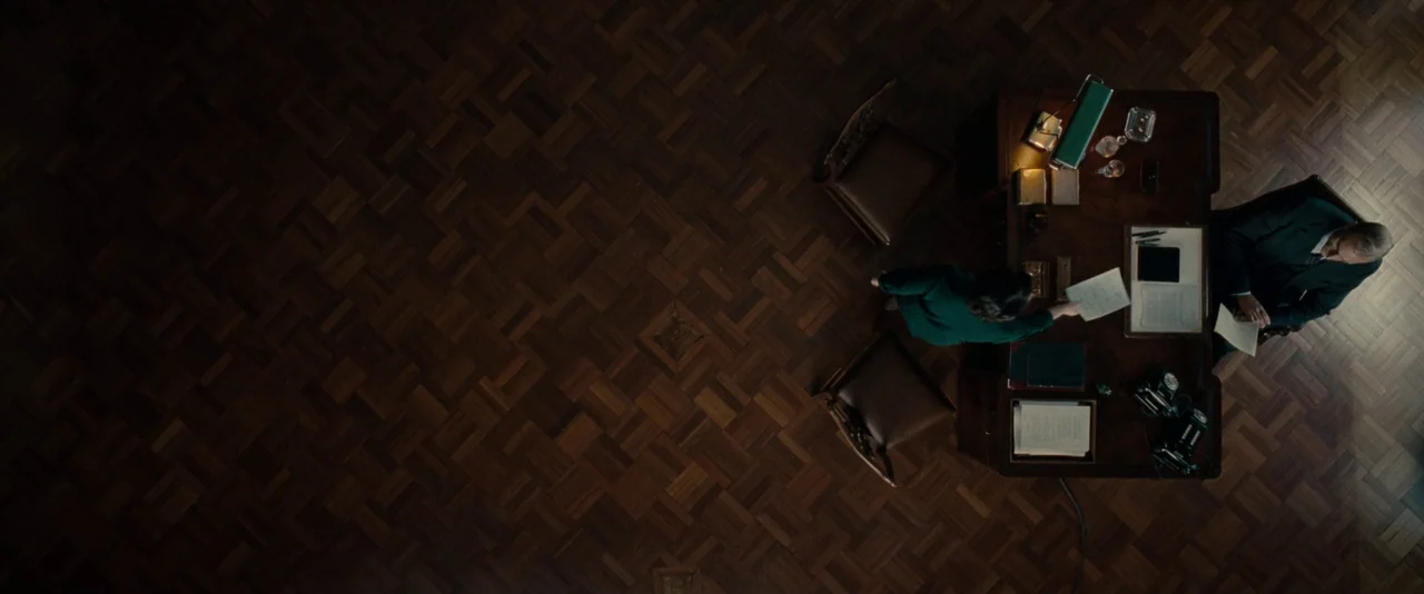 The Imitation Game (2014) movie still: wide — A high-angle view shows a dark, formal workspace with patterned wooden flooring. Two people interact…