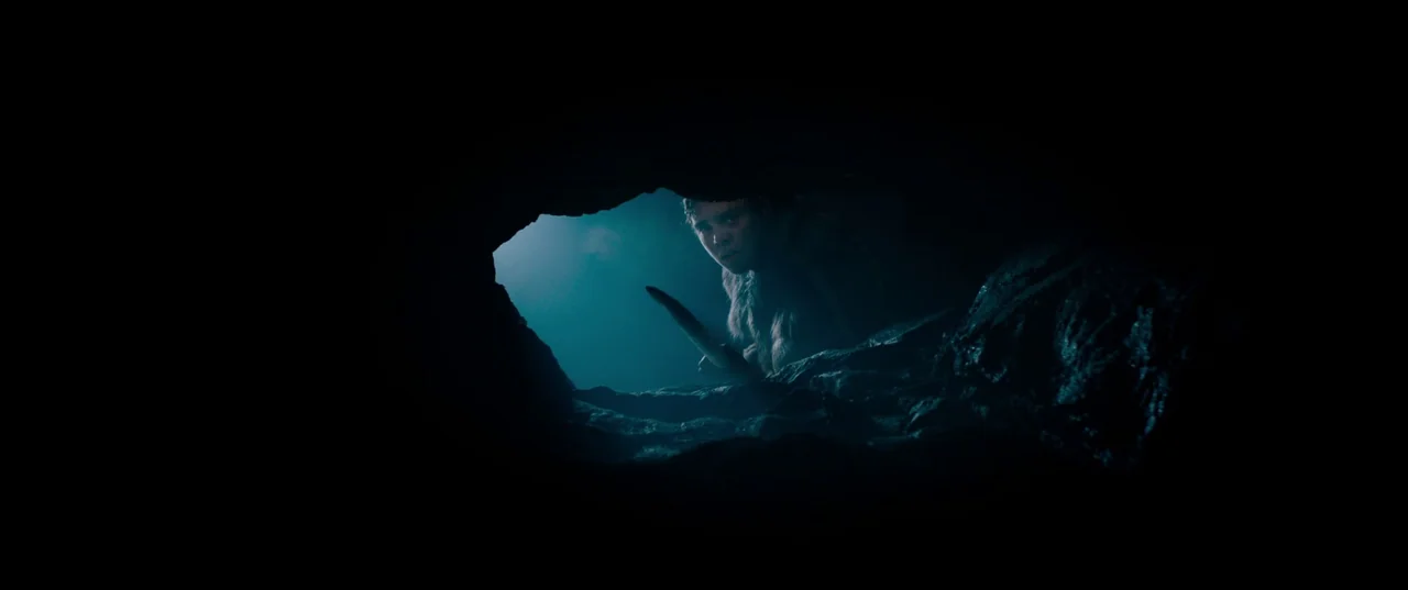 Out of Darkness (2022) movie still: wide — A dark, cramped interior of a rock cave is punctuated by a narrow horizontal opening. A young person in…