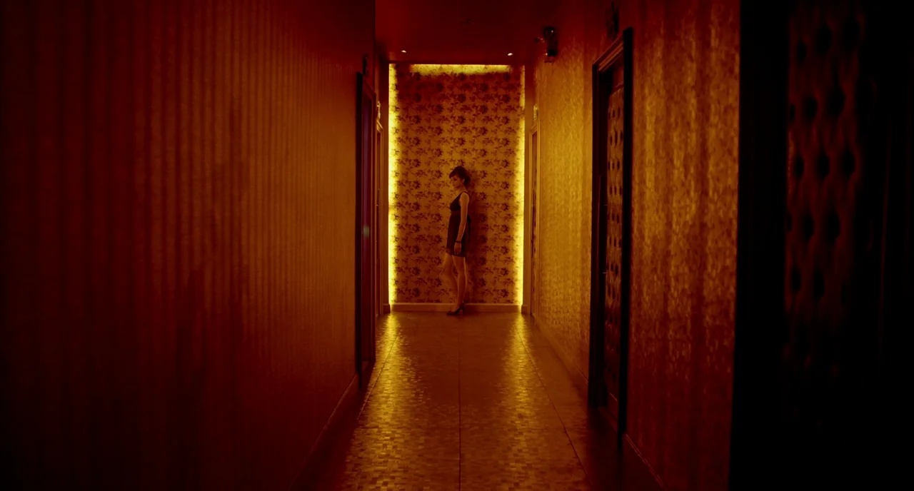 Only God Forgives (2013) movie still: wide — A narrow hotel corridor features textured dark walls and a tiled floor. A woman stands against a…