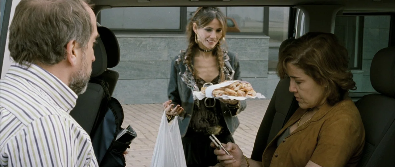 Rivals (1972) movie still: medium — The open cargo area of a car parked outdoors in a paved area. A woman holding a plate of pastries stands outside…