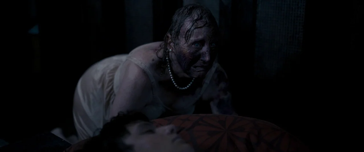 Talk To Me (2022) movie still: medium — A dark bedroom interior with a sleeping person in the foreground. An older woman with damaged skin crawls…