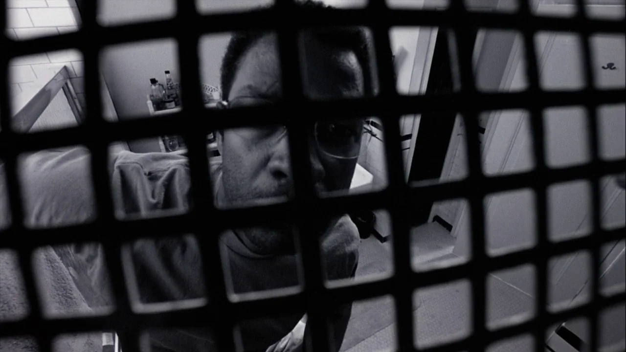 The Manchurian Candidate (2004) movie still: closeup — A man looks down from above through a dark, grid-patterned grate. The background shows tiled…