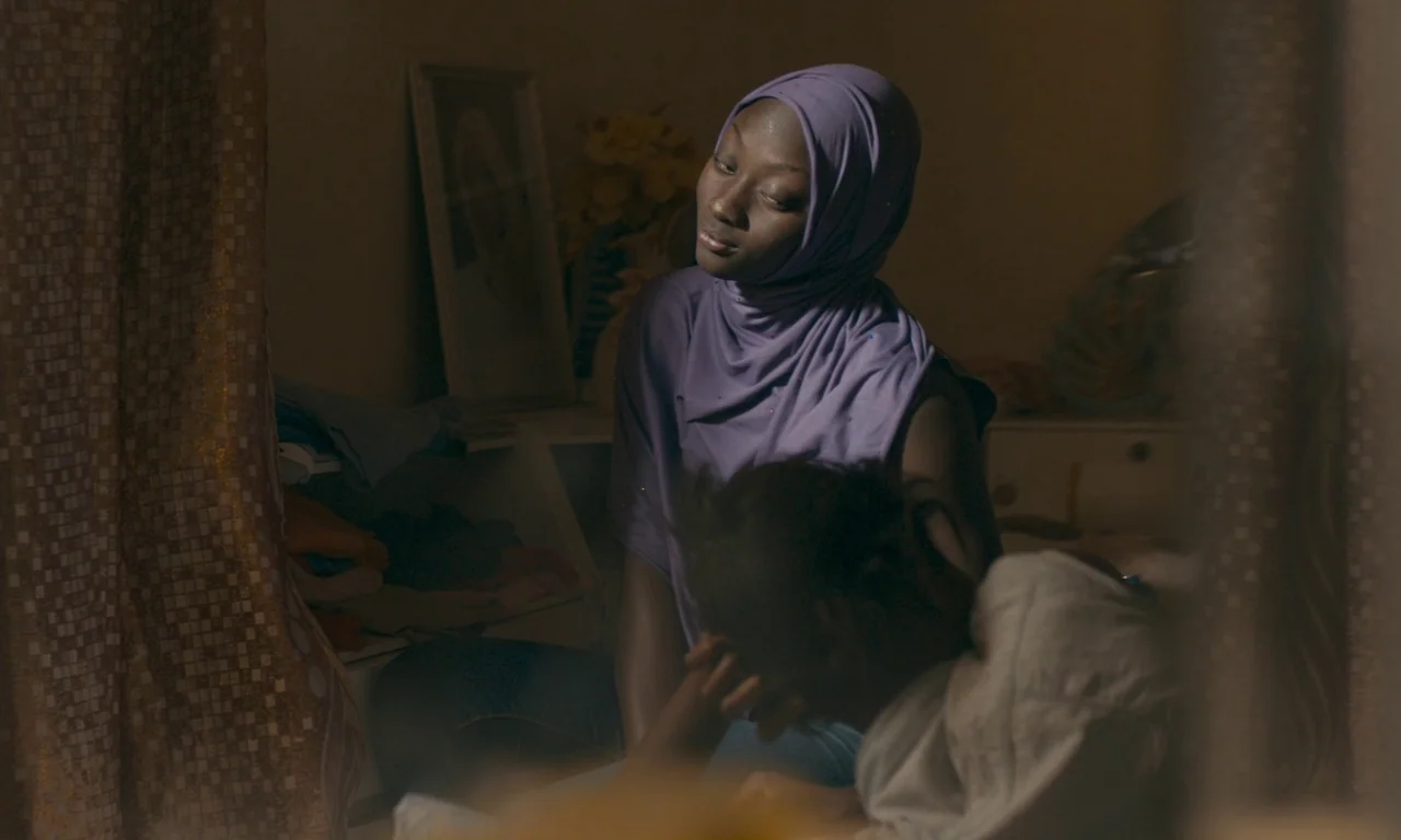 Atlantics (2019) movie still: medium — A dimly lit room features a woman sitting and looking toward a small child. The child rests their head on the…