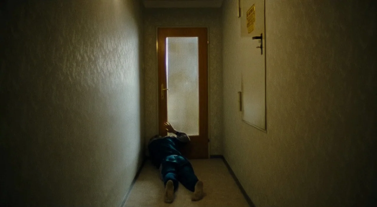 Paradise: Faith (2012) movie still: wide — A narrow, dimly lit residential hallway with plain textured walls and light carpet. A person lies prone…