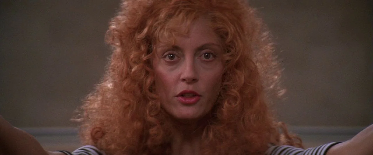 The Witches of Eastwick (1987) movie still: closeup — A woman with voluminous red hair stands in a studio against a plain backdrop. She faces…