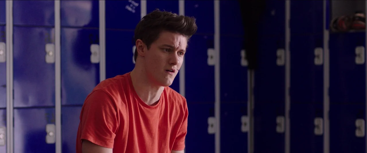 Anna and the Apocalypse (2018) movie still: medium — A row of glossy blue lockers lines the wall behind a young man. The man stands facing away from…