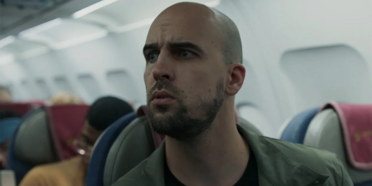 Fight or Flight (2025) movie still: medium — The interior of an airplane cabin features rows of passenger seats with maroon headrests. A bald man in…