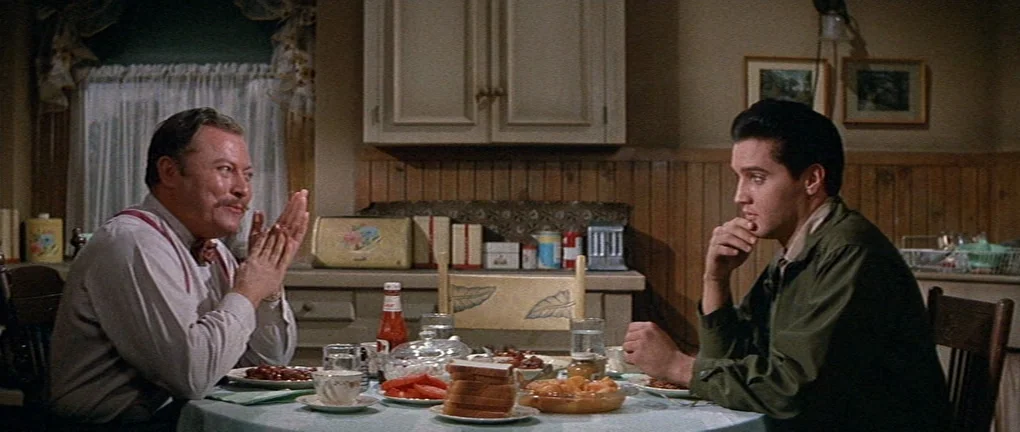 Wild in the Country (1961) movie still: medium — A 1960s domestic setting features wooden paneling, white cabinets, and a table with food. Two men…