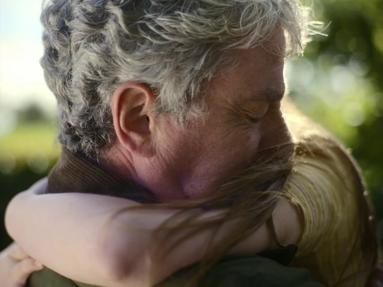 The Quiet Girl (2022) movie still: closeup — A man with silver hair and a child embrace in an outdoor setting. The sun illuminates the man's hair…