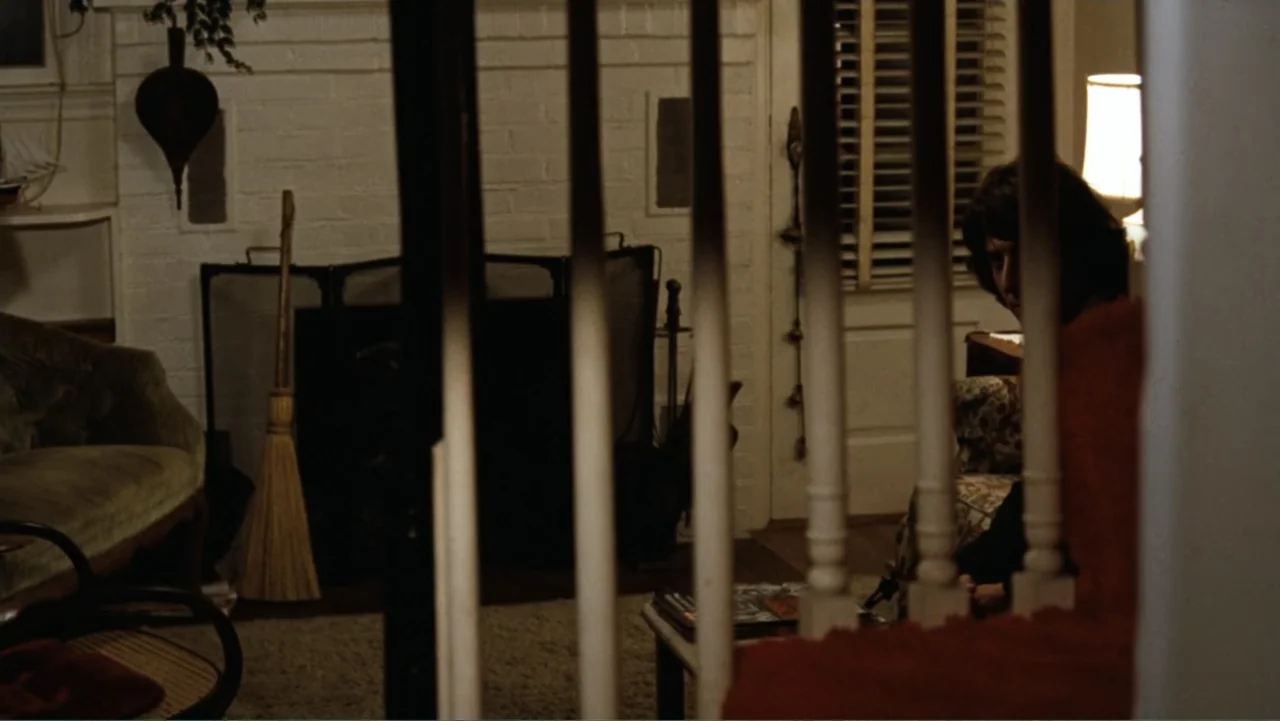 All the President's Men (1976) movie still: medium — A dark living room with a white painted brick fireplace and wood staircase railings. A man sits…