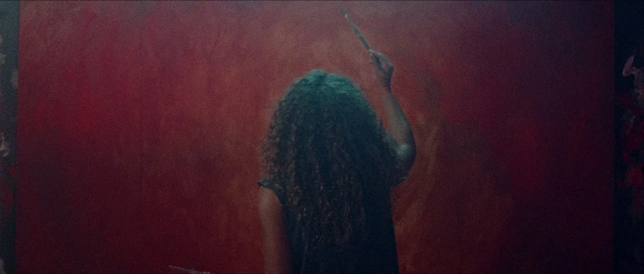 Bliss movie still: medium — A large, textured red canvas fills most of the frame in a dim room. A woman with long curly hair stands with her back to…