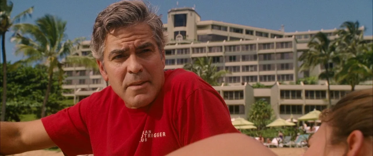 The Descendants (2011) movie still: medium — A man stands on a beach in front of a multi-story resort complex. Palm trees and beach umbrellas are…