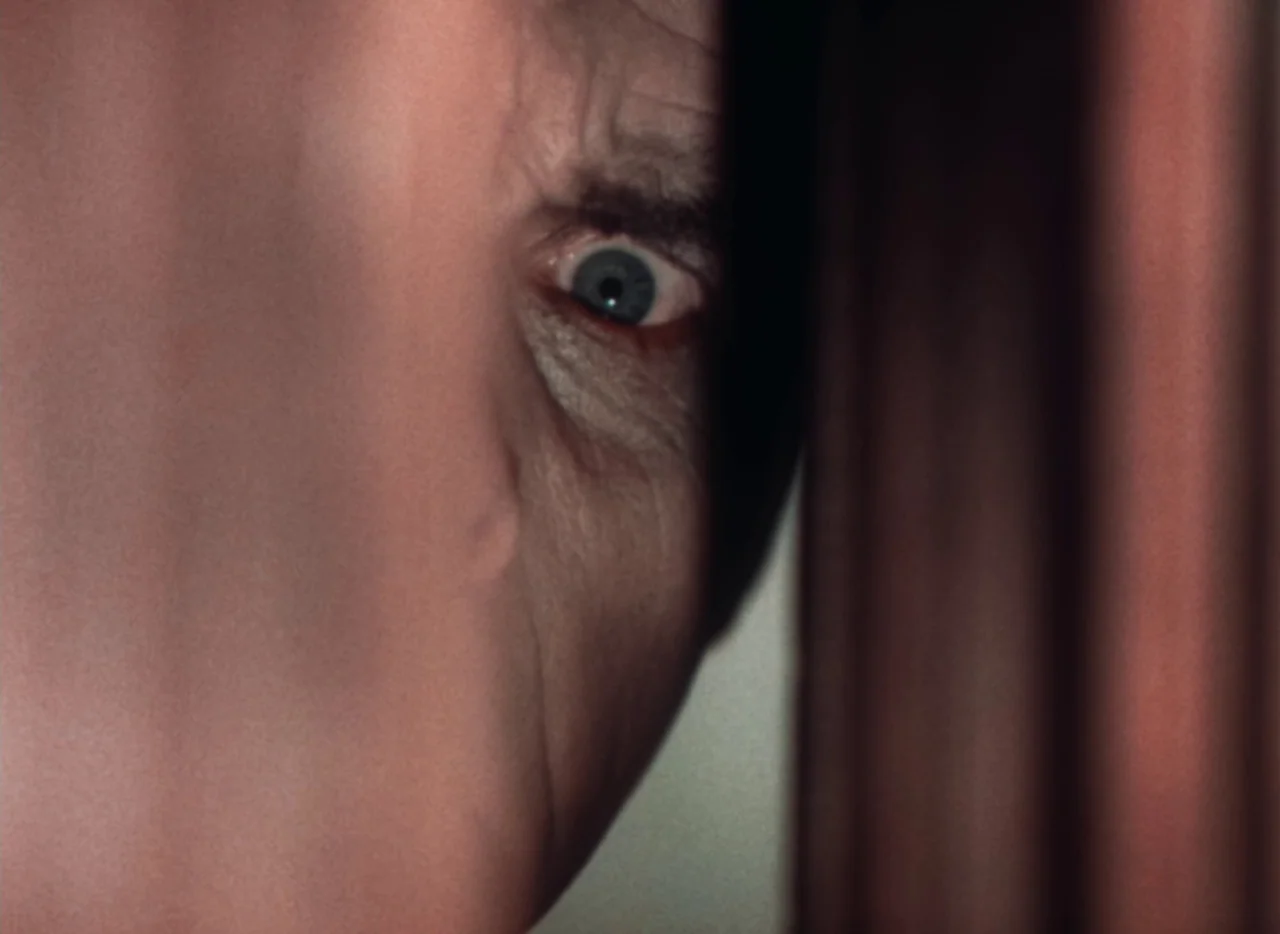 Doctor X (1932) movie still: closeup — A dark, narrow vertical slit between two heavy, brown fabric curtains. A single human eye stares directly…