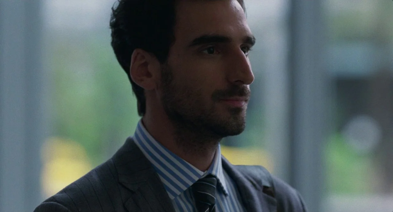Matthias & Maxime (2019) movie still: medium — A man in a professional business suit stands inside a modern office space. He looks toward the side…