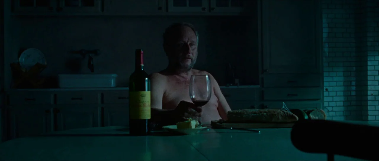 Inexorable (2021) movie still: medium — A dimly lit room with dark cabinets and a white tiled wall in background. A shirtless elderly man sits at a…
