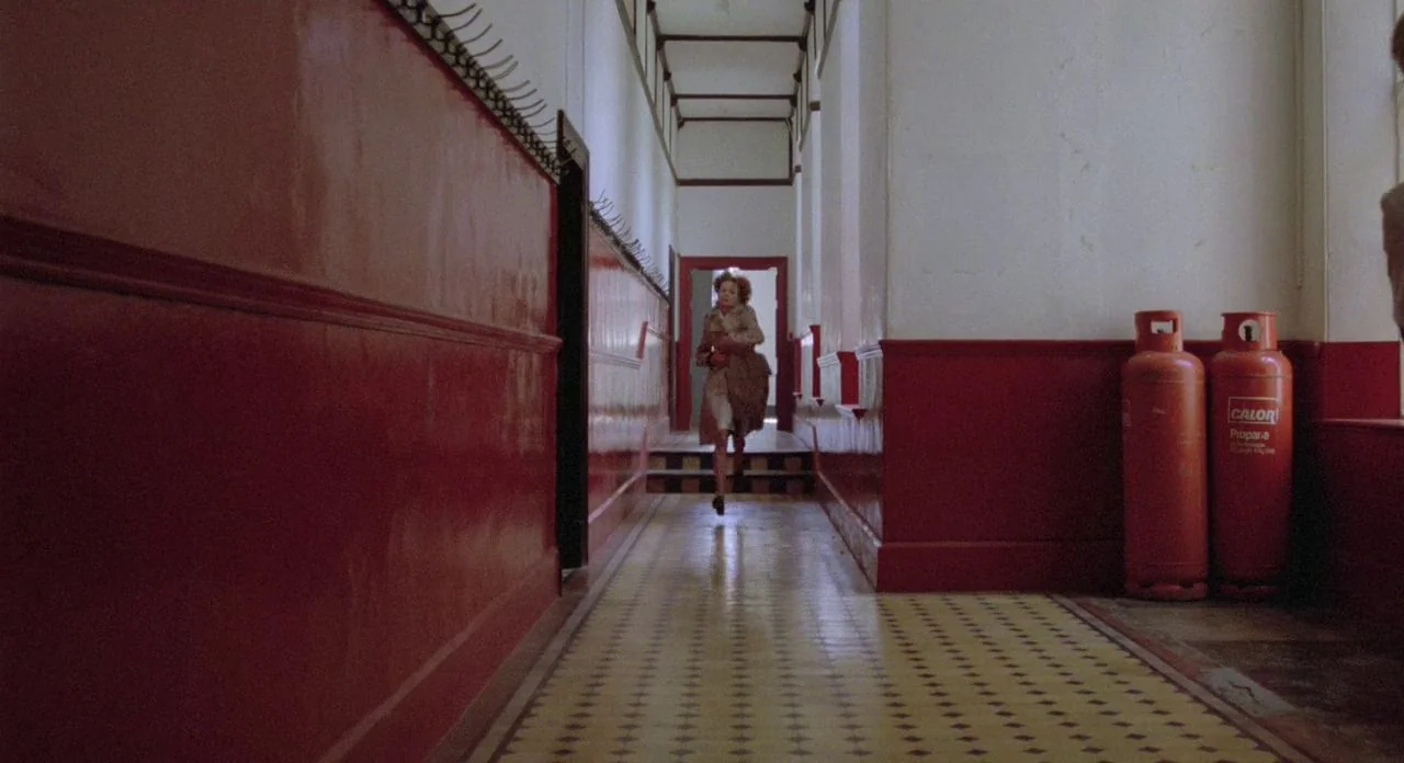 The Shout (1978) movie still: wide — A long corridor with glossy deep-red walls, white ceilings, and yellow-patterned tile flooring. A woman in a…