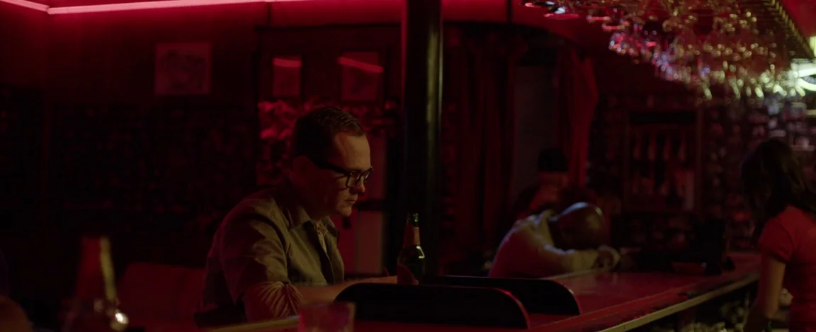 Cheap Thrills (2013) movie still: medium — A dimly lit drinking establishment features red neon lighting and a cluttered wooden surface. A man with…
