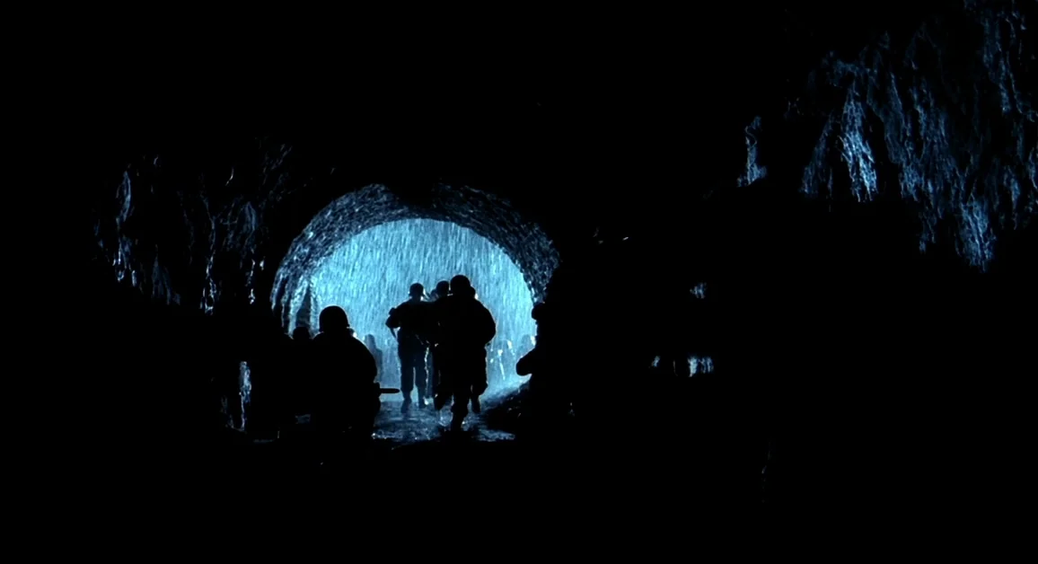 Hellboy (2004) movie still: wide — A dark stone tunnel frames a group of soldiers walking toward a stormy exit. Heavy rain falls outside the tunnel…