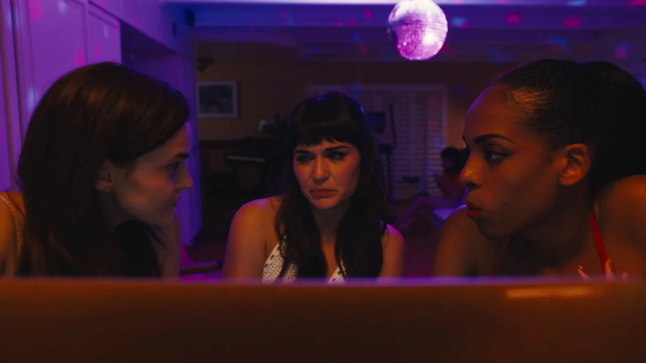 Cam (2018) movie still: medium — A dark, dimly lit room is illuminated by the light from a laptop screen. Three young women huddle around the…