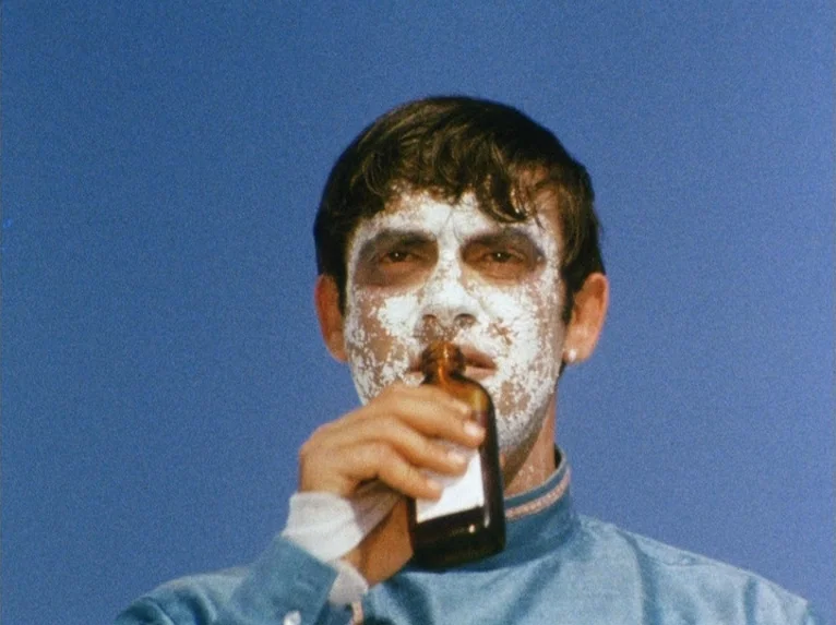 Our Lady of the Turks (1968) movie still: closeup — A man stands against a clear, solid blue sky background. He holds a small amber glass bottle to…