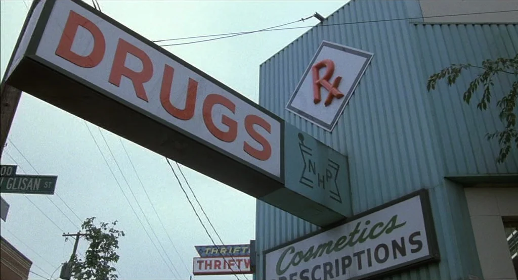 Drugstore Cowboy (1989) movie still: establishing — An exterior view of a pharmacy building features a prominent large hanging drugs sign. The…