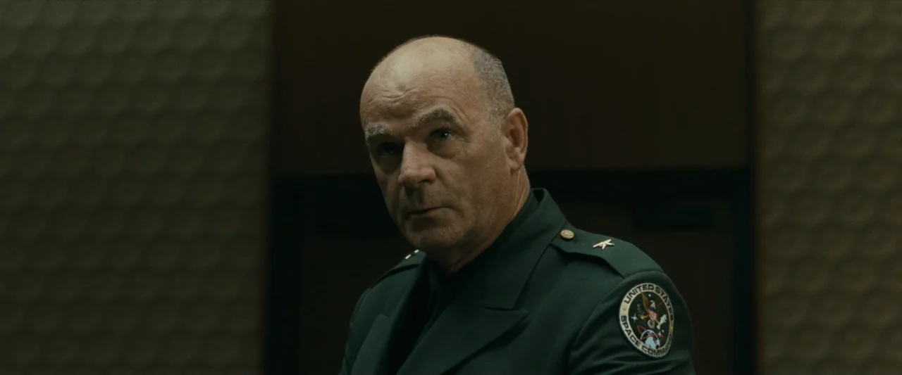 Ad Astra (2019) movie still: closeup — A dimly lit room with textured dark wall panels behind a military officer. A middle-aged man in a dark green…