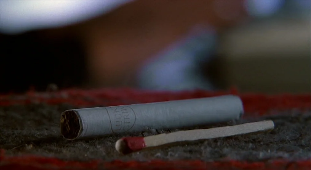 Misery (1990) movie still: closeup — A white Lucky Strike cigarette and a wooden match lie on a rough surface. The objects are presented in a static…