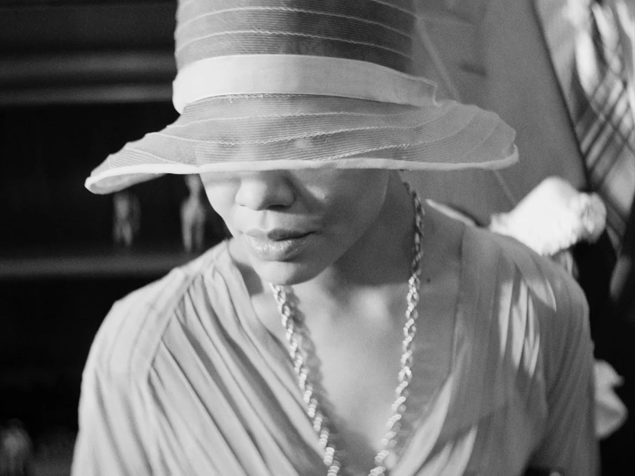 Passing (2021) movie still: closeup — An interior space contains a woman wearing a 1930s style wide-brimmed hat. She looks downward while the hat…