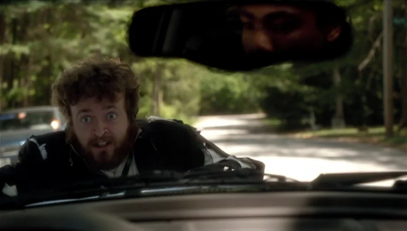 Mystery Team (2009) movie still: medium — A man with curly hair and a beard leans over a car hood on a road. A second person is visible within the…