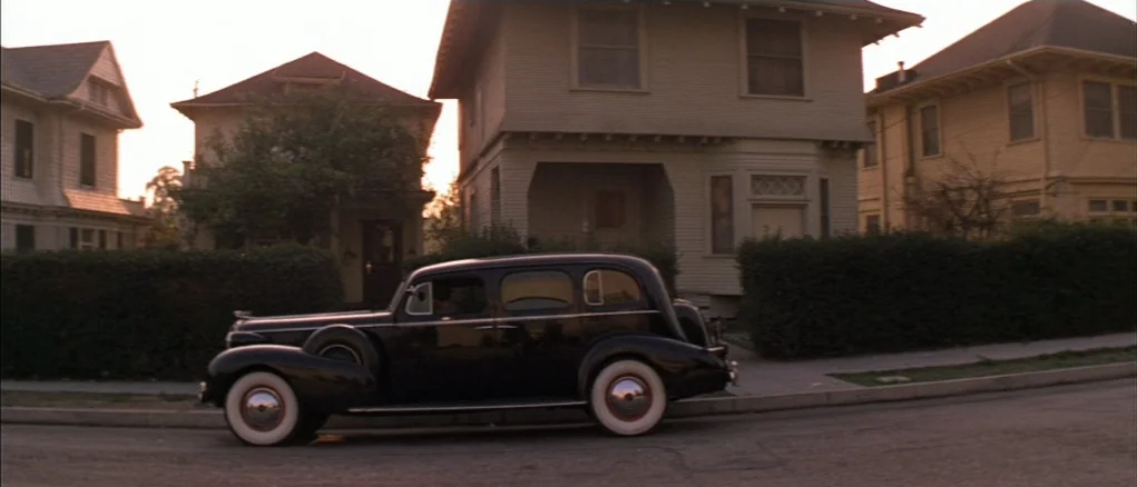 Chinatown (1974) movie still: wide — A quiet residential street lined with multi-story, 1930s-style American suburban homes at sunset. A dark…