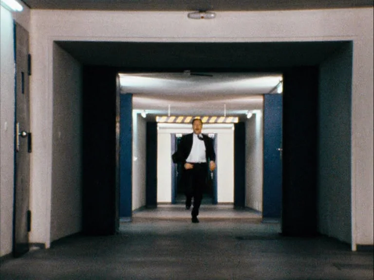 Making of: World On A Wire (1973) movie still: wide — A long, sterile industrial hallway features white walls and dark blue accents. A man in a…