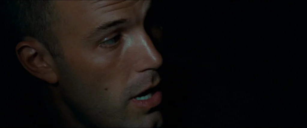 The Town (2010) movie still: closeup — A man's face is illuminated against a solid black background. He is speaking while turned slightly away from…