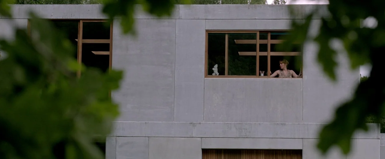 Borgman (2013) movie still: wide — A modern concrete building features large rectangular windows framed in wood. A woman stands inside the…
