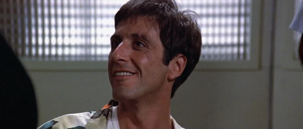 Scarface (1983) movie still: closeup — The subject sits inside a room with a white grid-patterned window treatment behind him. The man has a slight…