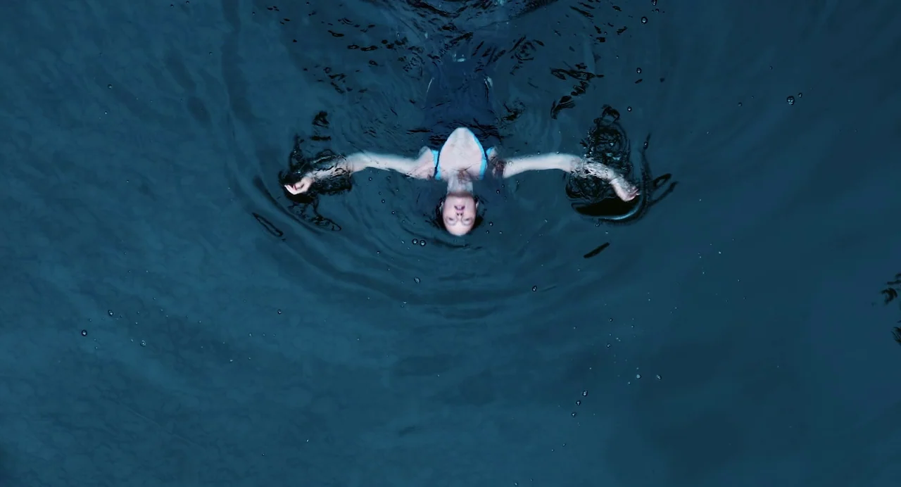 Hellbender (2021) movie still: wide — A person floats on their back in the center of dark, expansive open water. The camera captures them from a…