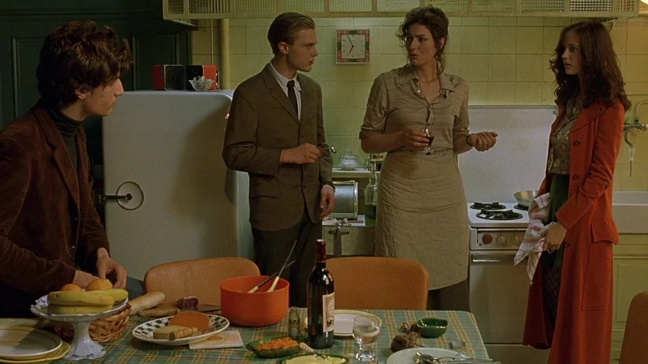 The Dreamers (2003) movie still: medium — A 1960s European kitchen features green tile walls and a checkered table surface. Four people gather…