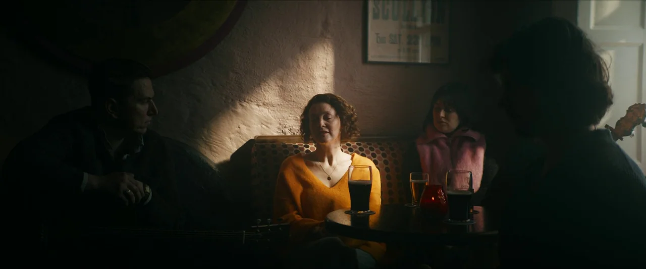 Kneecap (2024) movie still: medium — A dimly lit pub booth with patterned fabric seating and dark wood tables. Three people sit around a table with…