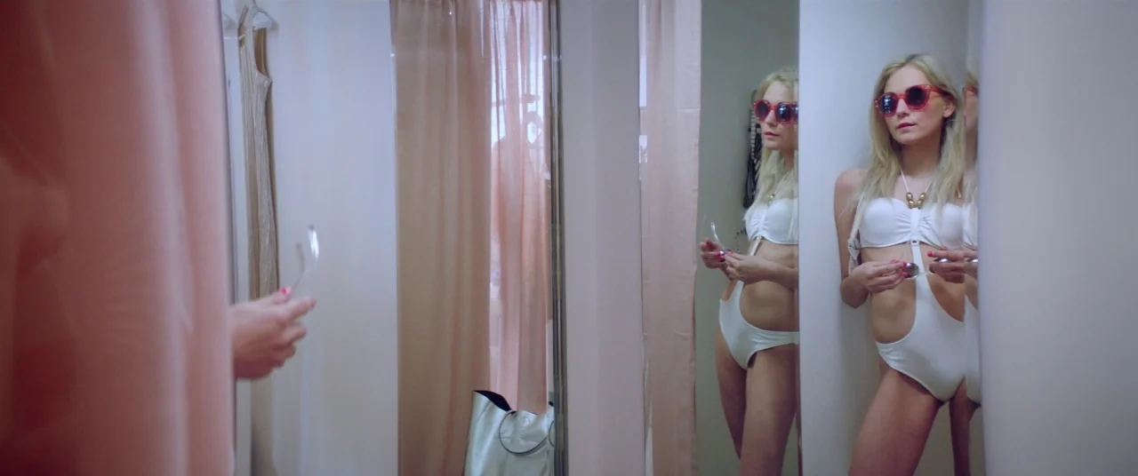Holiday movie still: medium — A cramped fitting room with white walls and sheer pale pink fabric curtains. A young blonde woman in a white swimsuit…