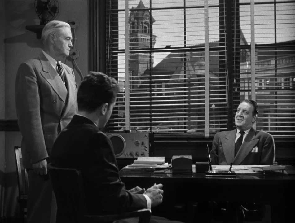 Strangers on a Train (1951) movie still: medium — An office with a desk and venetian blinds looking out onto a brick building. Two men stand and sit…