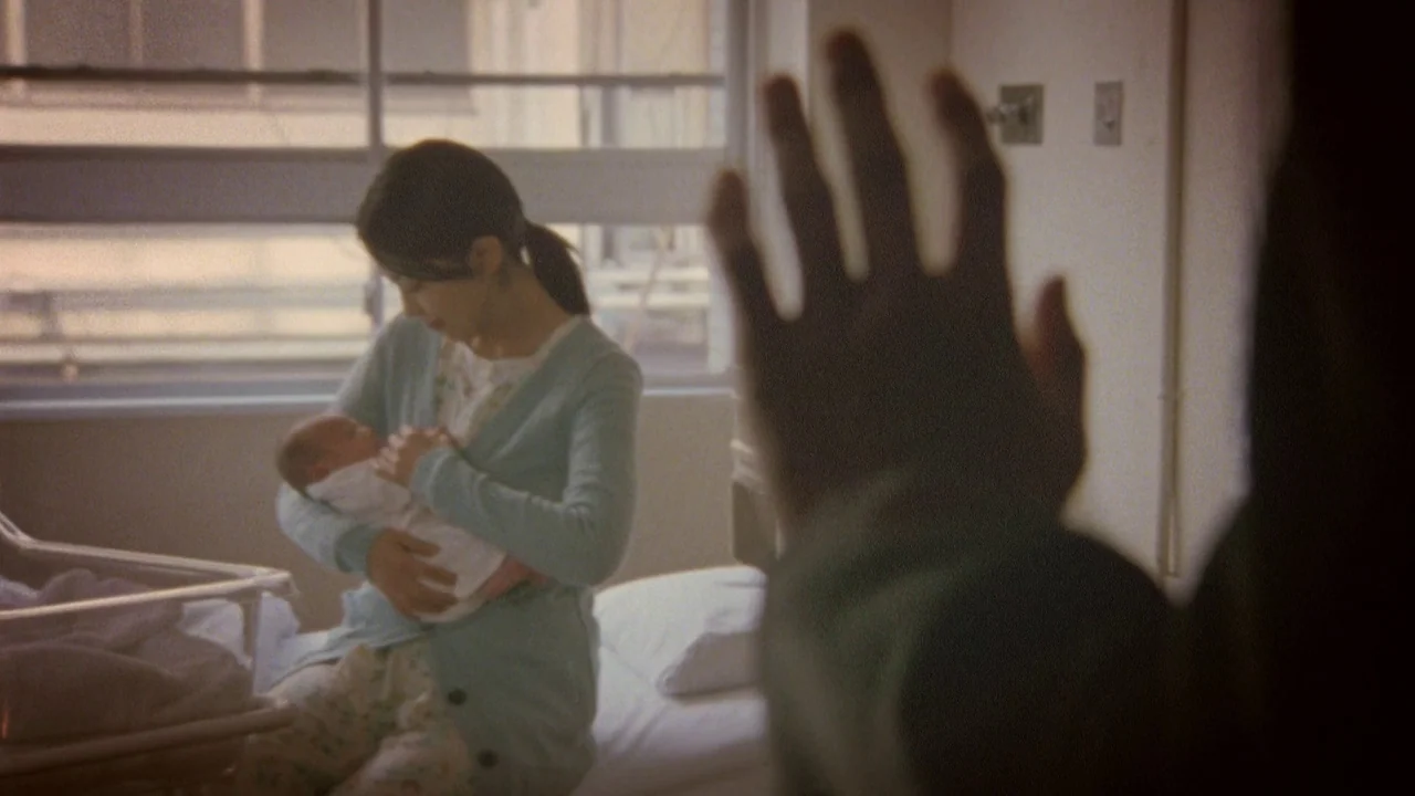 Confessions (2010) movie still: medium — A hospital room with a window wall, plain walls, and a medical bassinet nearby. A woman sits on the bed…