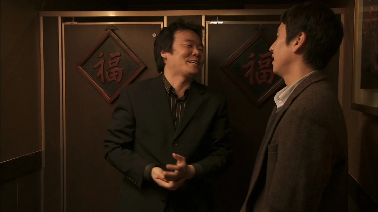 Oki’s Movie (2010) movie still: medium — An interior space features two men standing against wood-paneled walls adorned with red symbols. One man in…