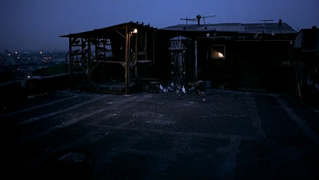 Ghost Dog: The Way of the Samurai (1999) movie still: establishing — A dark rooftop features a wooden pigeon coop illuminated by a single warm bulb.…