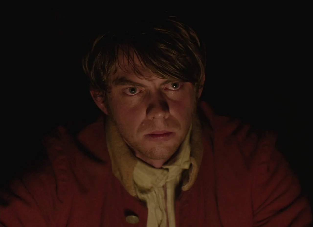 The Nightingale (2018) movie still: closeup — A dark outdoor space at night with no visible background details. A man in a red coat is illuminated…